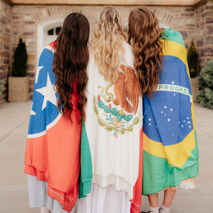 Three LDS missionaries standing together with personalized mission flag blankets draped over their shoulders, featuring Puerto Rico, Mexico, and Brazil flags.