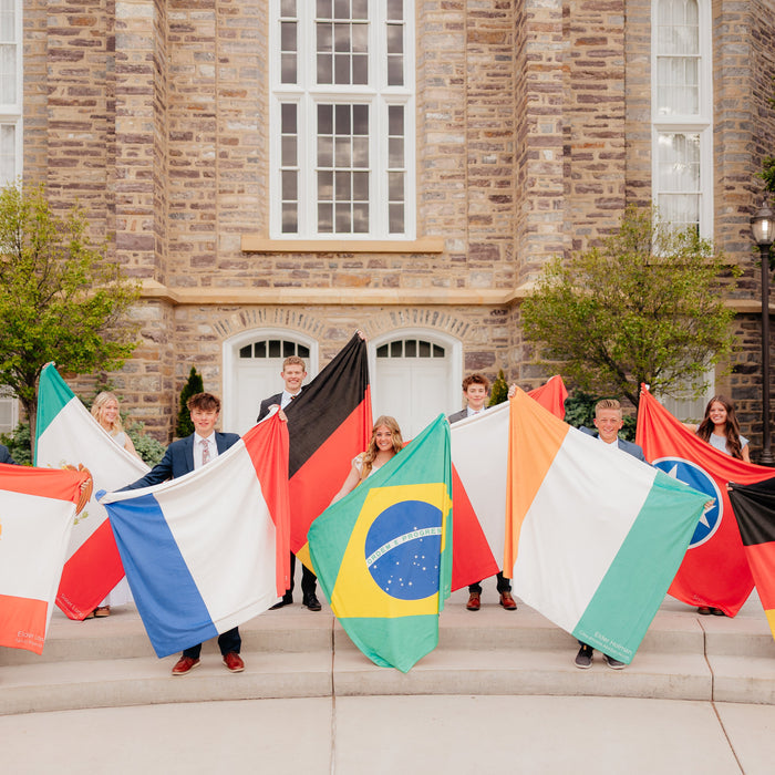 LDS missionaries holding personalized mission flag blankets in front of a temple, featuring custom country flags and missionary service locations.