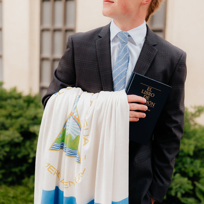 LDS missionary holding a personalized mission flag blanket and Book of Mormon outside a temple, celebrating missionary service and faith.