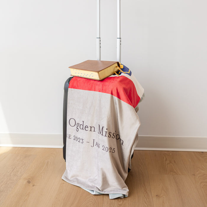 Personalized mission flag blanket draped over a missionary suitcase with scriptures on top, prepared for LDS missionary service.