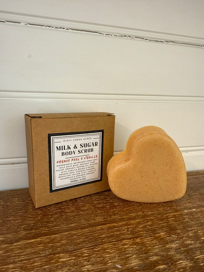 Heart-shaped body scrub next to its packaging on a wooden surface with a white wall background.