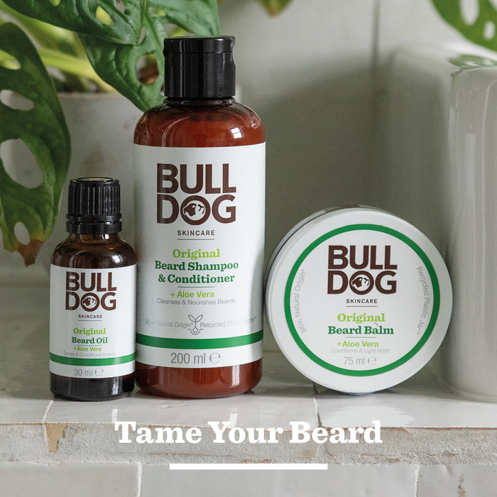 Bulldog Skincare’s Original Beard Oil, made with natural ingredients, is displayed on a surface with green plants in the background. Text below reads, “Tame Your Beard.”.