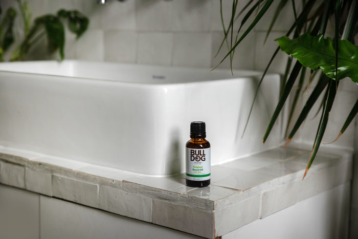 A brown bottle of Original Beard Oil with natural ingredients sits on the tiled edge of a white bathroom sink, surrounded by green leafy plants.