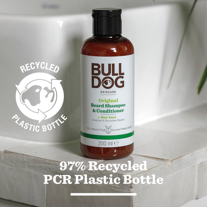 A bottle of Original Beard Shampoo and Conditioner rests on a bathroom counter, its label featuring natural origin ingredients and eco-friendly packaging made from 97% recycled PCR plastic, plus a visible recycling symbol.