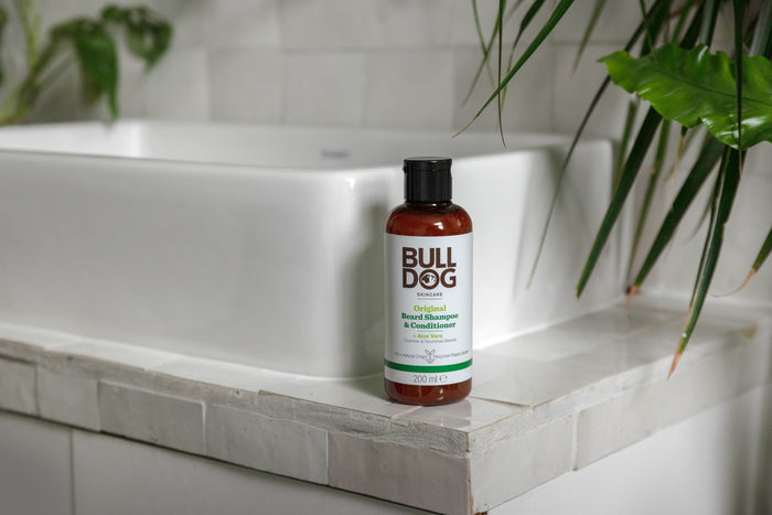 A bottle of Original Beard Shampoo and Conditioner, made with natural origin ingredients, sits on the edge of a white tiled bathroom sink with green plants nearby.