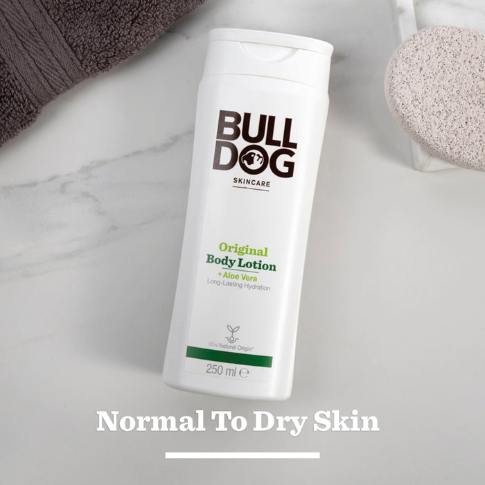 A white bottle of Original Body Lotion is placed on a marble surface beside a brown towel and a round exfoliating sponge. Normal To Dry Skin appears at the bottom of the image.