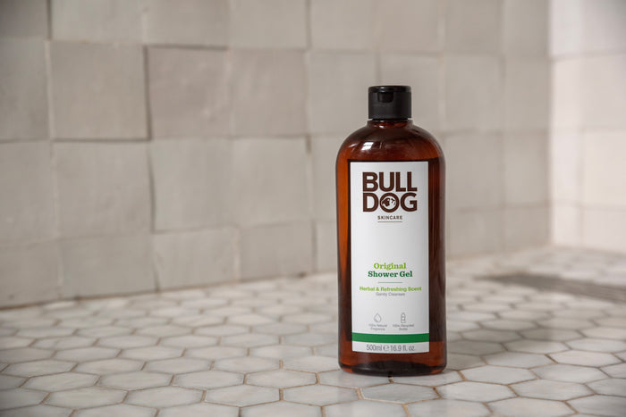 A bottle of Original Shower Gel stands on a tiled bathroom floor with gray and white hexagonal tiles and a beige tiled wall in the background, offering a refreshing scent with natural ingredients.