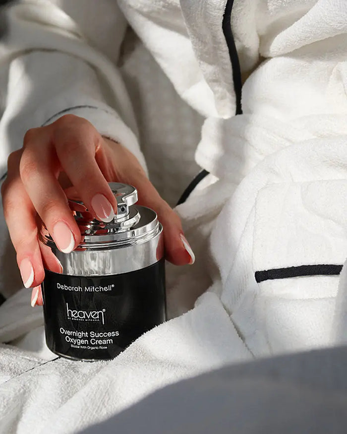 Hand holding a black and silver canister labeled 'Deborah Mitchell Heaven' against a white background