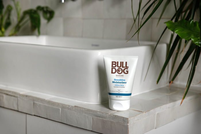 A tube of Sensitive Moisturiser rests on the edge of a white tiled bathroom sink, with green plants visible in the background.