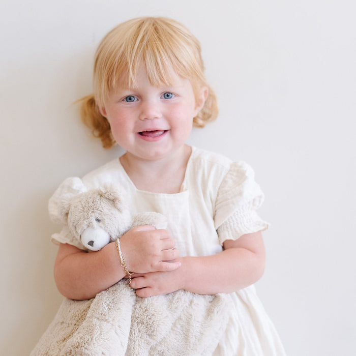 Adorable toddler girl in a white dress cuddling a Saranoni bear stuffed animal lovey. This ultra-soft plush lovey blanket features a cozy bear head and luxuriously soft fabric, designed to provide comfort, security, and soothing snuggles for babies and toddlers. Made with high-quality materials, the Saranoni lovey is perfect for newborns, infants, and young children as a bedtime companion or travel comfort item. Ideal baby shower gift or nursery essential, offering warmth and softness in a neutr