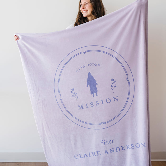 Woman holding a personalized missionary blanket featuring the Utah Ogden Mission design, missionary name, and custom mission dates on a soft lavender blanket.