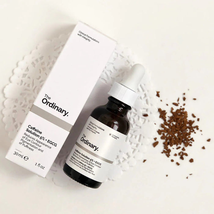 The Ordinary skincare serum bottle and packaging on a textured white surface with scattered coffee beans.