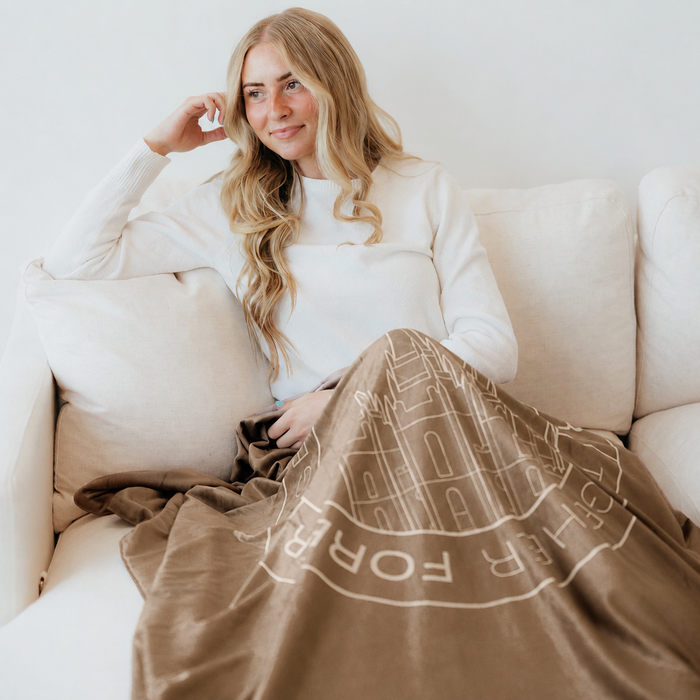 Woman relaxing on a couch wrapped in a cocoa brown LDS blanket featuring a temple design and the words "Together Forever," soft faith-inspired throw blanket celebrating eternal families and lasting connections.