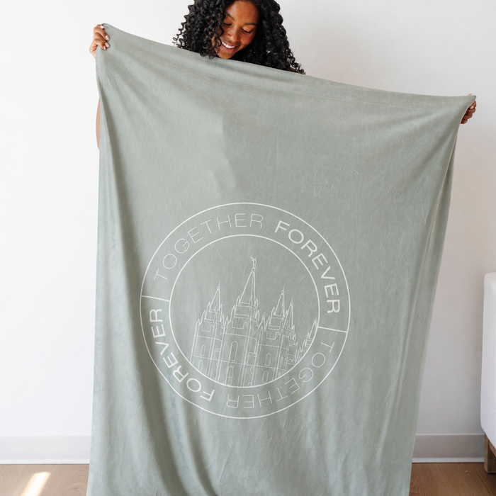 Person holding a light olive green LDS blanket featuring a temple design and the words "Together Forever," soft faith-inspired throw blanket celebrating eternal families and lasting connections.