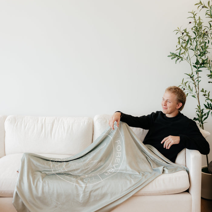 Person relaxing on a couch wrapped in a light olive green LDS blanket featuring a temple design and the words "Together Forever," soft faith-inspired throw blanket celebrating eternal families and lasting connections.
