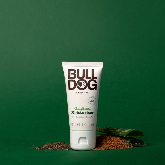 A tube of Travel Original Moisturiser stands upright on a green background, surrounded by aloe vera, seeds, and green tea leaves, emphasizing its natural ingredients. The white tube features black and green text.