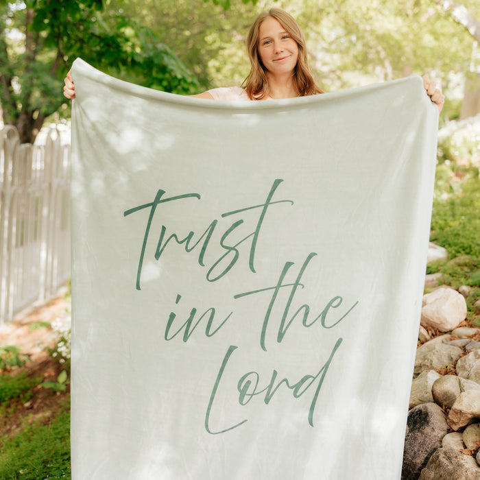 Woman holding a light rosemary green Christian blanket featuring the words "Trust in the Lord" outdoors, soft faith-inspired throw blanket for comfort, encouragement, and reflection.