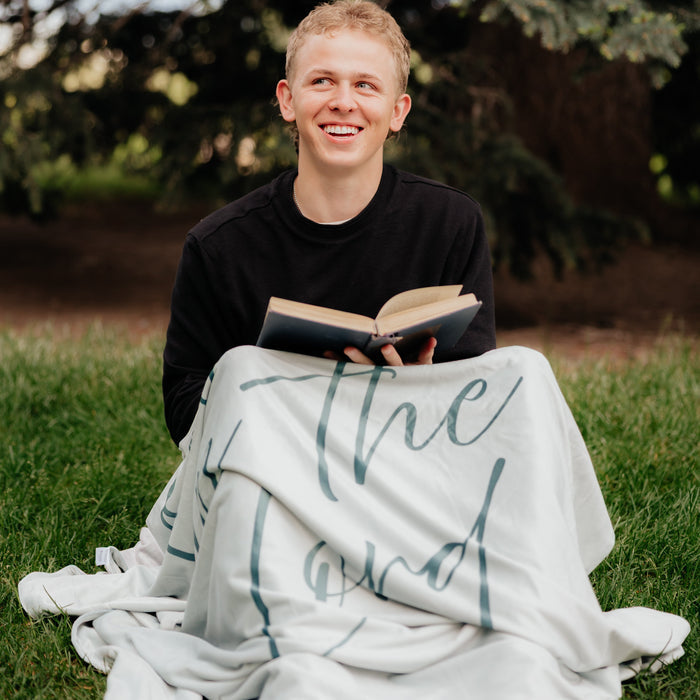 Person reading a Bible outdoors while wrapped in a light rosemary green Christian blanket featuring the words "Trust in the Lord," faith-inspired throw blanket for comfort, encouragement, and reflection.