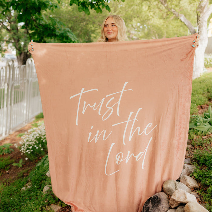 Woman holding a camel brown Christian blanket featuring the words "Trust in the Lord" outdoors, soft faith-inspired throw blanket for comfort, encouragement, and peace.