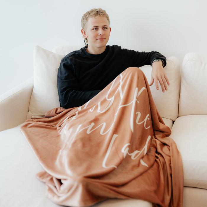 Person relaxing on a couch wrapped in a camel brown Christian blanket featuring the words "Trust in the Lord," soft faith-inspired throw blanket for comfort, encouragement, and peace.