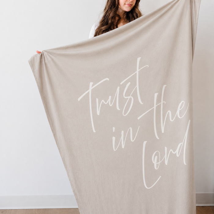 Person holding a light cobblestone gray Christian blanket featuring the words "Trust in the Lord," soft faith-inspired throw blanket displayed against a neutral indoor background.