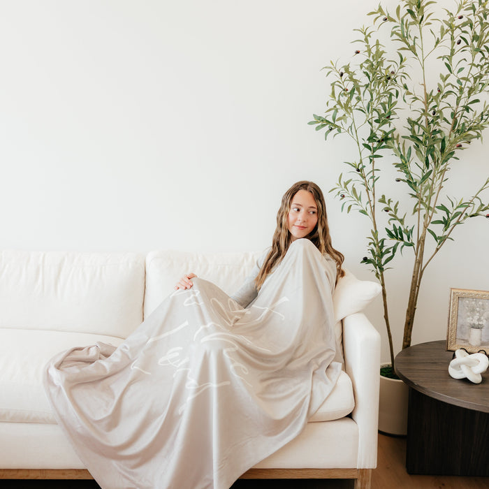 Woman relaxing on a couch wrapped in a light cobblestone gray Christian blanket featuring the words "Trust in the Lord," soft faith-inspired throw blanket for comfort, encouragement, and peace.