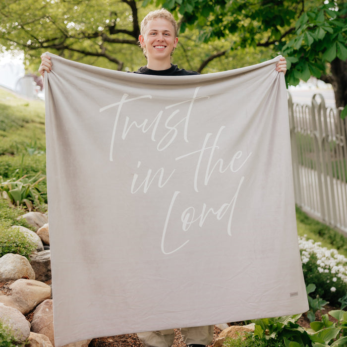 Person holding a light cobblestone gray Christian blanket featuring the words "Trust in the Lord" outdoors, soft faith-inspired throw blanket for comfort, encouragement, and peace.