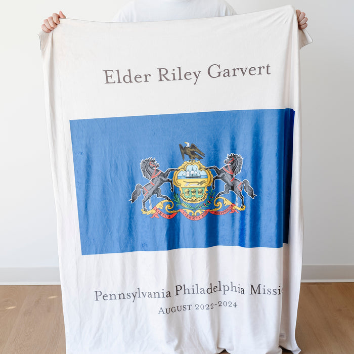 Person holding a personalized missionary blanket featuring the Pennsylvania Philadelphia Mission flag, missionary name, and custom mission dates.