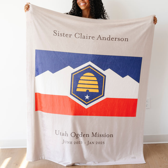 Woman holding a personalized missionary blanket featuring the Utah Ogden Mission flag, missionary name, and custom mission dates.