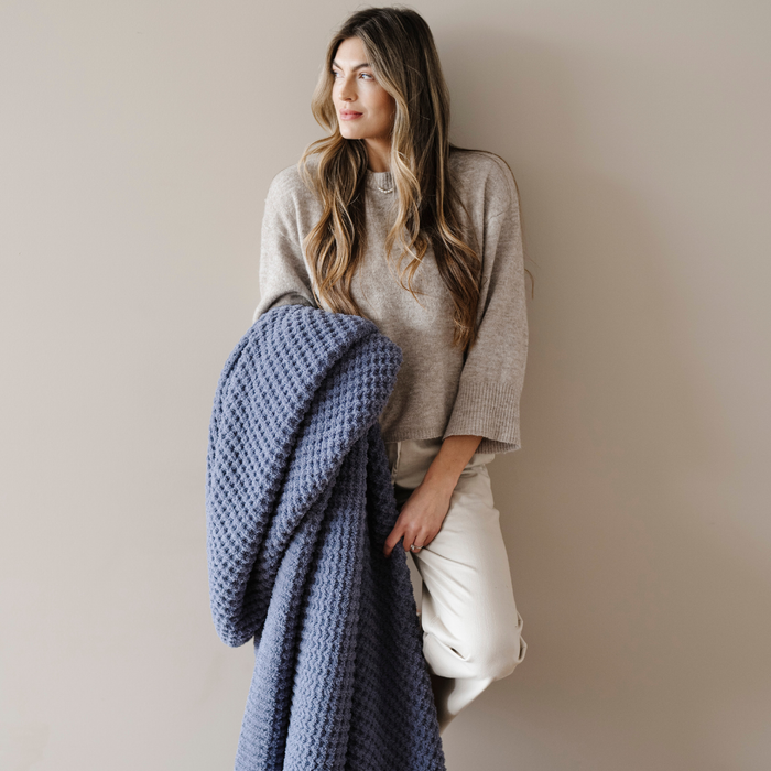 A woman is holding a blue waffle knit blanket. This luxury blanket is very soft and is a Saranoni blanket. Use this cozy blanket for relaxing, gifting, or as a home decor piece. Embrace luxury that lasts and enjoy this throw blanket for years to come!