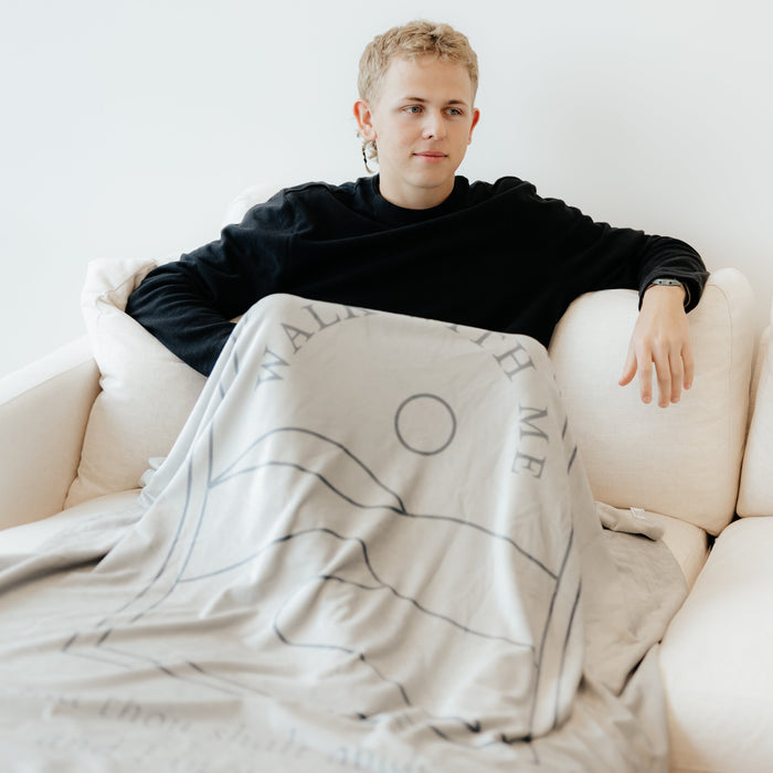 Young man sitting on a couch wrapped in an ash gray LDS youth theme blanket featuring the words "Walk With Me," faith-centered throw blanket inspired by the 2026 LDS Youth Theme and a reminder to walk with Jesus Christ.