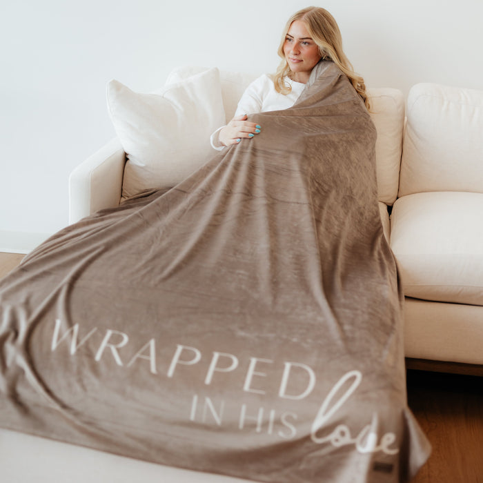 Woman wrapped in a soft cocoa brown Christian blanket featuring the words "Wrapped in His Love" while relaxing on a couch.