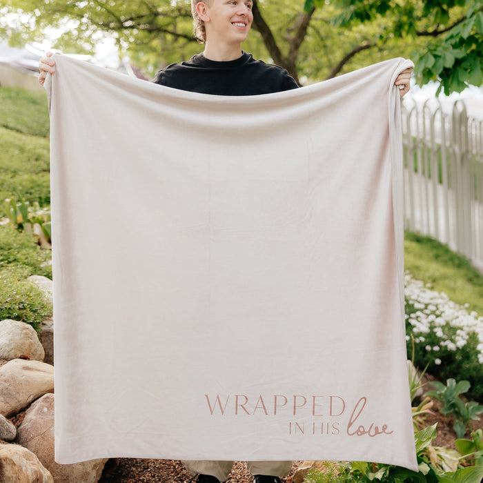 Person holding a cream Christian blanket featuring the words "Wrapped in His Love" in a peaceful outdoor setting.