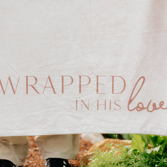 Close-up of a cream Christian blanket featuring the words "Wrapped in His Love" in elegant script lettering.
