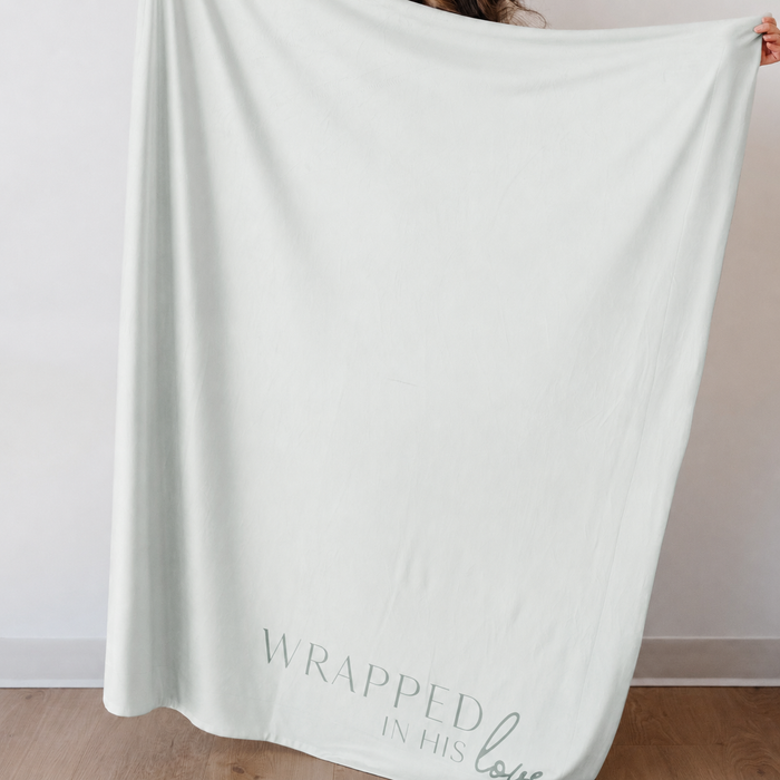 Light green Christian blanket featuring the phrase "Wrapped in His Love" displayed by a person, faith-inspired throw blanket gift for comfort and encouragement.