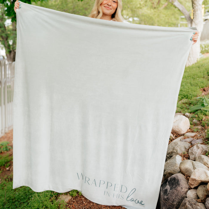 Person holding a light green Christian blanket featuring the phrase "Wrapped in His Love" outdoors, soft faith-inspired throw blanket for comfort and encouragement.